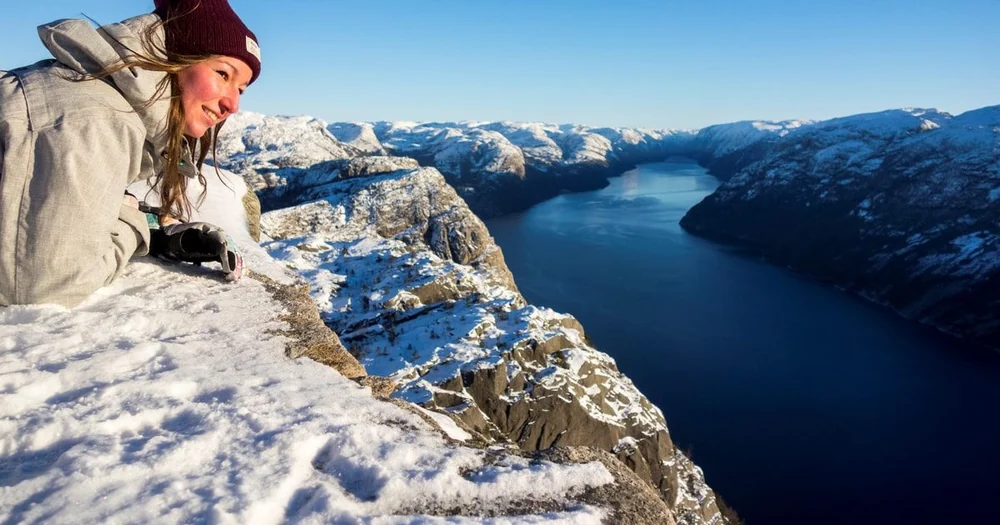 Eine lächelnde Frau mit Mütze, die am Klippenrand im Schnee liegt und auf einen langen blauen Fjord zwischen schneebedeckten Bergen hinabblickt